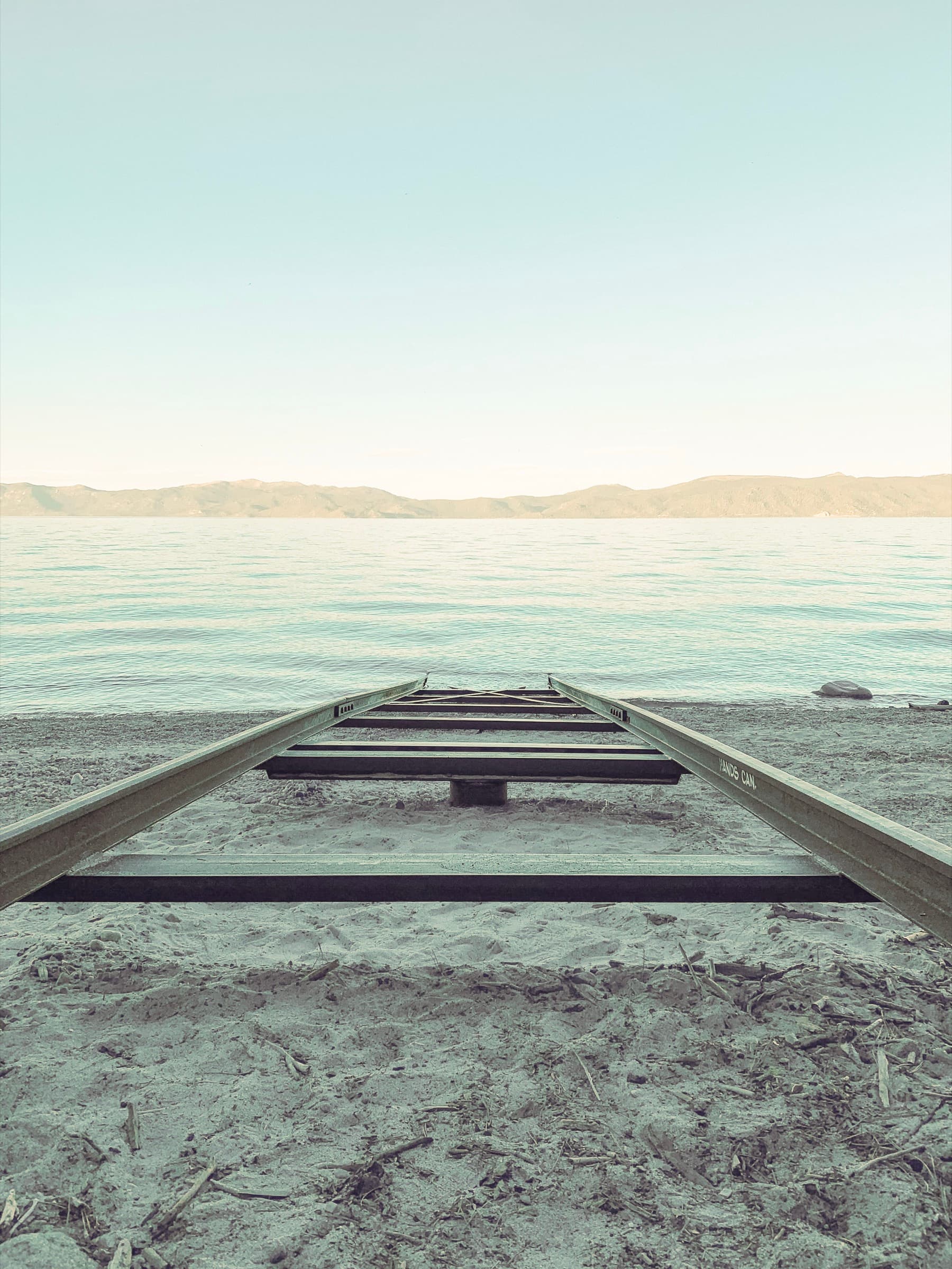 A metal boat-launch rail receding from a sandy shore into the calm water of Lake Tahoe, mountains on the far shore beneath a wide pale sky.
