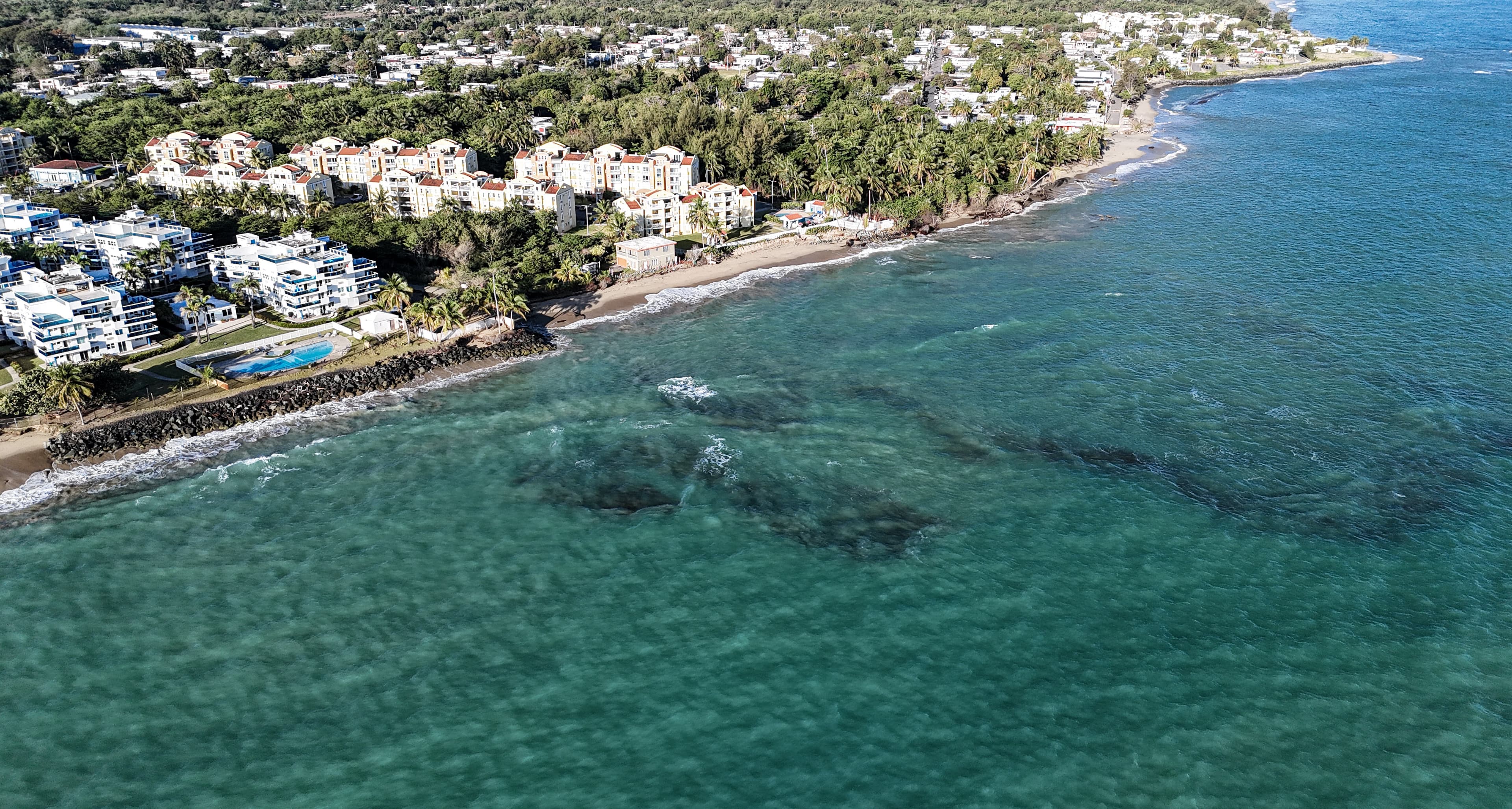 A drone view of a Puerto Rican coastline: turquoise reef water, a pale sand beach, dense palms, and white condominiums with tiled roofs.